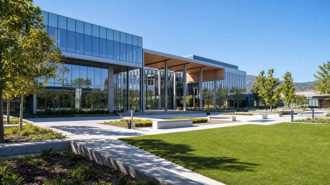 Architekturaufnahme eines modernen Technologiecampus mit Glasfassade, gepflegten Rasenflächen und öffentlichen Plätzen unter kalifornischem Sonnenlicht – repräsentiert die Büroinfrastruktur von Meta Platforms