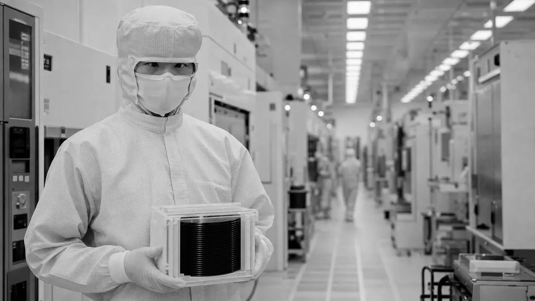 Schwarz-Weiß-Reportagefoto eines Reinraum-Technikers im weißen Bunny-Suit, der einen Wafer-Carrier vor Maschinenreihen hält