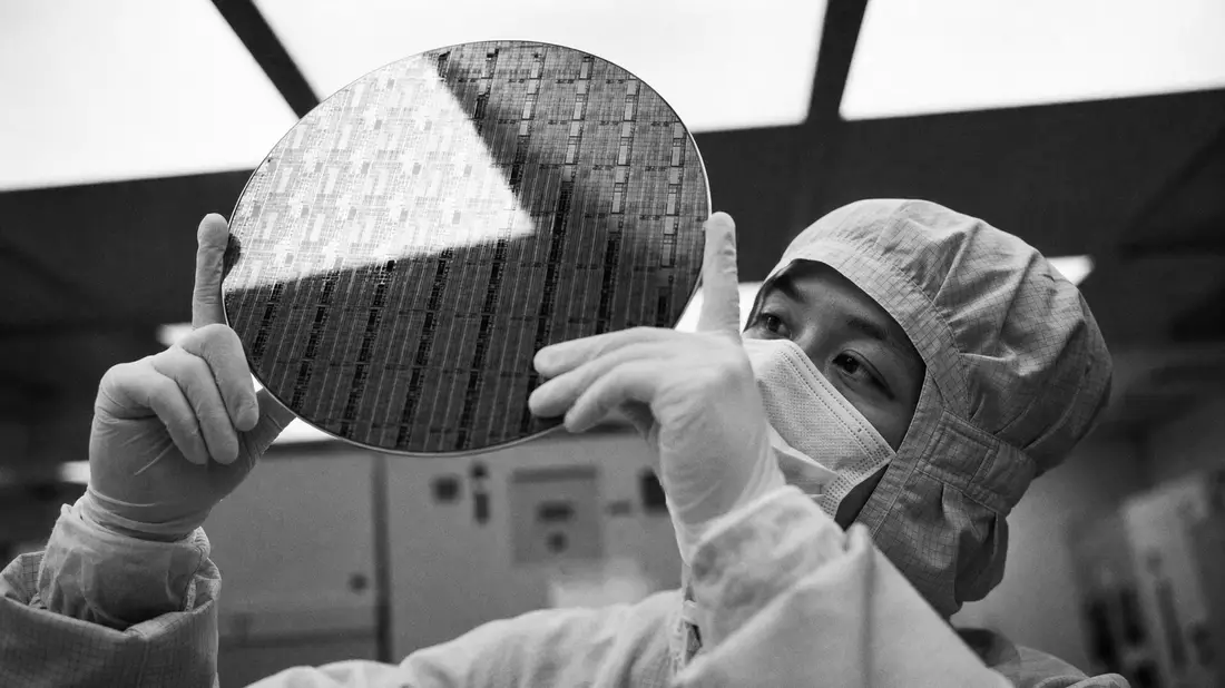 Schwarz-Weiß-Reportagefoto eines Halbleitertechnikers im Reinraumanzug, der einen großen NAND-Flash-Siliziumwafer gegen das Licht hält, dramatischer Kontrast