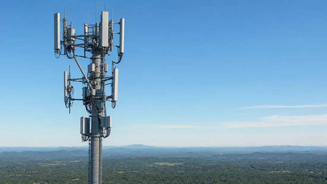 Hoher Funkmast mit 5G-Antennenpanels vor blauem Himmel und grüner Hügellandschaft