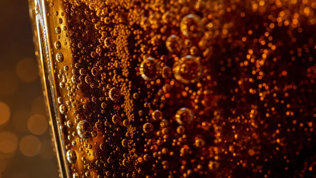Extremes Makrofoto von Kohlensäureblasen in einem dunklen generischen Getränk im Glas, goldenes Bernsteinlicht bricht sich in der Flüssigkeit, extreme Tiefenunschärfe