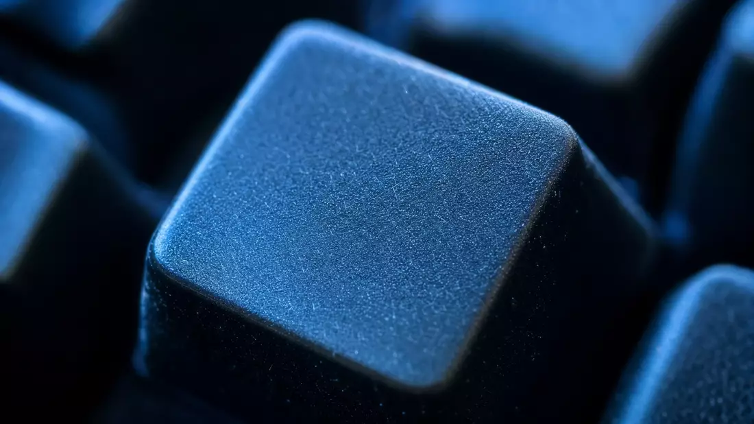 Extreme Makroaufnahme einer leeren Computertaste mit detaillierter Oberflächenstruktur im blauen Monitorlicht, starke Tiefenunschärfe lässt benachbarte Tasten verschwimmen, keine Beschriftung sichtbar