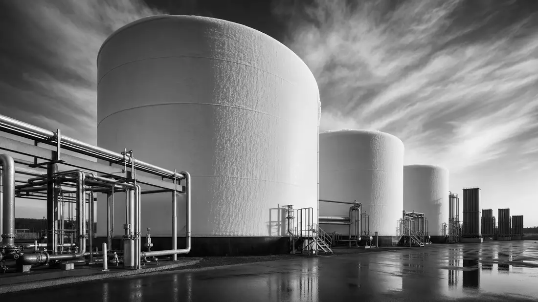 Schwarz-Weiß-Reportagefoto von drei massiven zylindrischen Kryotanks mit vereister Oberfläche, dramatischen Wolken und einem Netz aus Industrierohren im Vordergrund