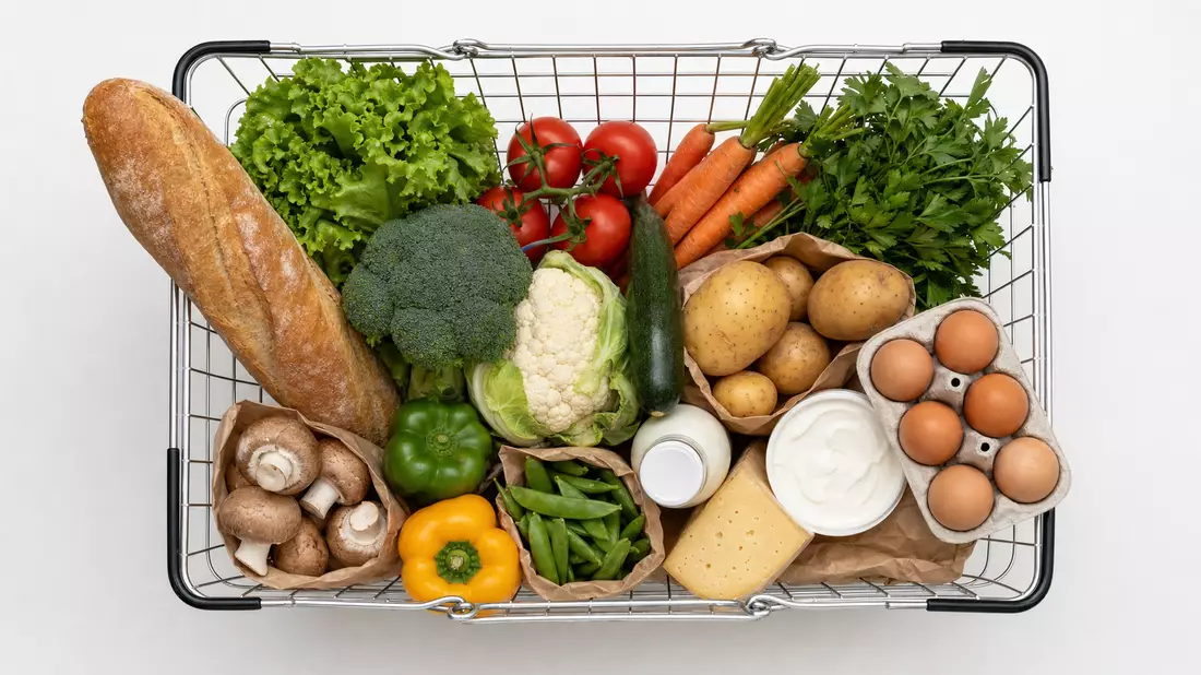 Flatlay-Draufsicht auf einen generischen Metalleinkaufskorb, gefüllt mit frischen Lebensmitteln: Gemüse, Baguette, Eier, Milch und Käse – sauber arrangiert auf weißem Hintergrund