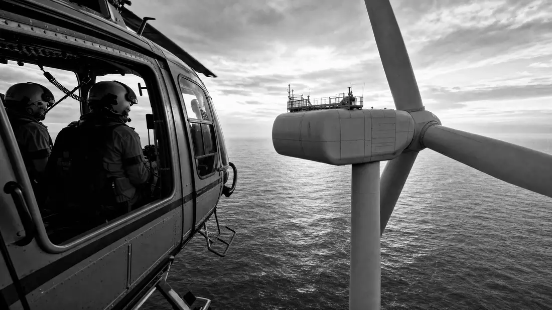 Black and white reportage photo of helicopter crew approaching an offshore wind turbine over the sea