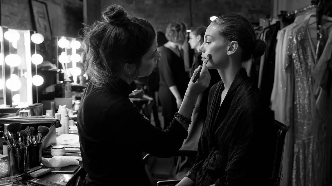 Black and white backstage reportage of a make-up artist applying foundation to a model at a fashion show