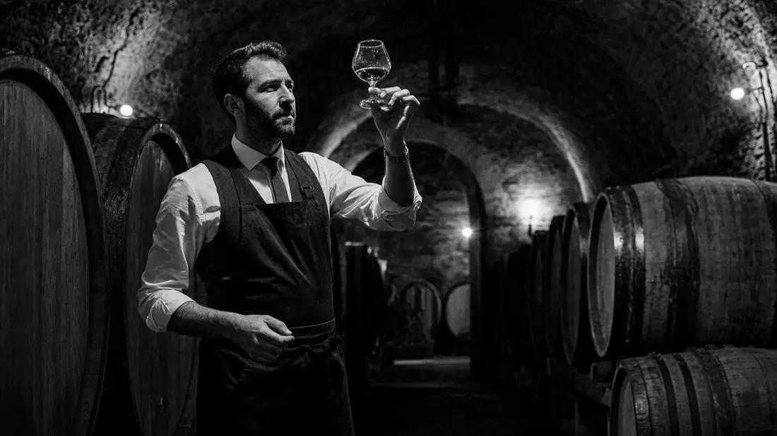 Black and white photo of sommelier examining cognac glass in vaulted oak barrel cellar
