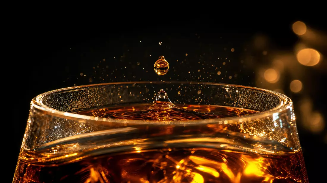 Extreme macro of a golden amber droplet frozen above a crystal whisky glass on black background