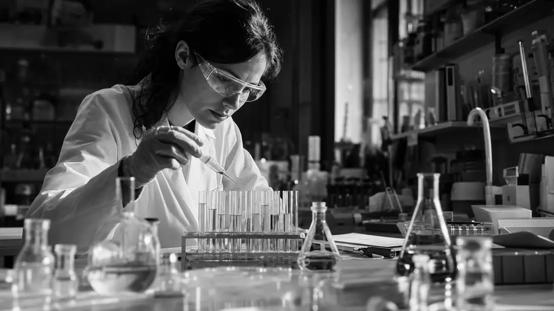 Black and white documentary photograph of a female scientist with safety goggles holding a pipette over test tubes in a research laboratory