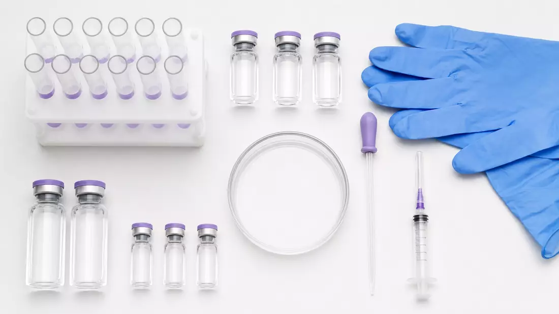 Overhead flatlay of pharmaceutical objects on white surface including clear glass vials, a syringe, petri dish, test tubes, a pipette, and blue nitrile gloves