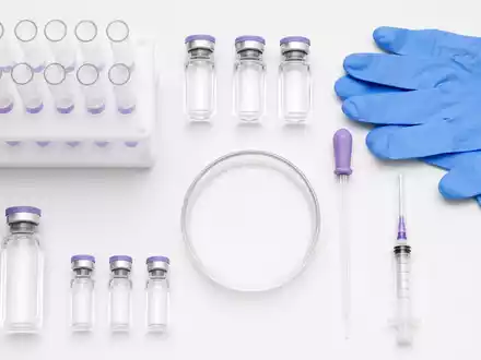 Sanofi FR0000120578 overhead flatlay of pharmaceutical vials, syringe, pipette, petri dish, and protective gloves - Foto: THN