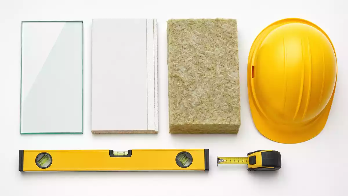Overhead flat lay of glass panel, plasterboard, mineral wool, spirit level, measuring tape and hard hat on white