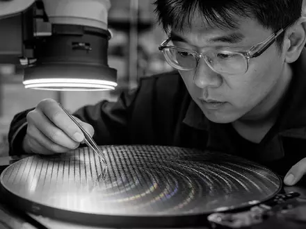 STMicroelectronics NL0000226223 engineer uses tweezers inspecting silicon wafer under bright microscope lamp - Foto: THN