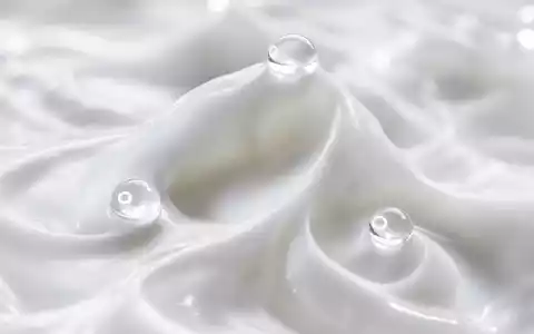 Extreme Makroaufnahme weißer Cremtextur mit Wellen und Spitzen; drei kristallklare Wassertropfen fangen das Licht. Flacher Schärfebereich, perlige Weißtöne, kein Verpackungsmaterial sichtbar - Foto: THN