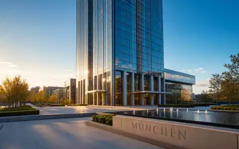Fotorealistisches Weitwinkel-Außenbild eines modernen Hochhaus-Firmensitzes in München bei goldenem Abendlicht. Glas-Stahl-Fassade spiegelt den Himmel, Plaza mit Wasserspiel vorn. Steinsockel-Inschrift „MÜNCHEN”. Allianz SE (DE0008404005) - Foto: THN