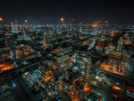 Cinematische Drohnenaufnahme eines weitläufigen Petrochemie-Komplexes bei Nacht aus ca. 300 Metern Höhe. Hunderte beleuchteter Rohrleitungen, Destillationskolonnen und Reaktortürme bilden ein leuchtendes Gitternetz. Bernsteinfarbene, weiß-blaue Industrielichter spiegeln sich auf dem Boden und in Dampfwolken. Anamorphe Linsenoptik, kinematisches Teal-Shadow-Grading - Foto: THN