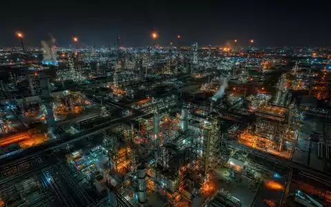 Cinematische Drohnenaufnahme eines weitläufigen Petrochemie-Komplexes bei Nacht aus ca. 300 Metern Höhe. Hunderte beleuchteter Rohrleitungen, Destillationskolonnen und Reaktortürme bilden ein leuchtendes Gitternetz. Bernsteinfarbene, weiß-blaue Industrielichter spiegeln sich auf dem Boden und in Dampfwolken. Anamorphe Linsenoptik, kinematisches Teal-Shadow-Grading - Foto: THN