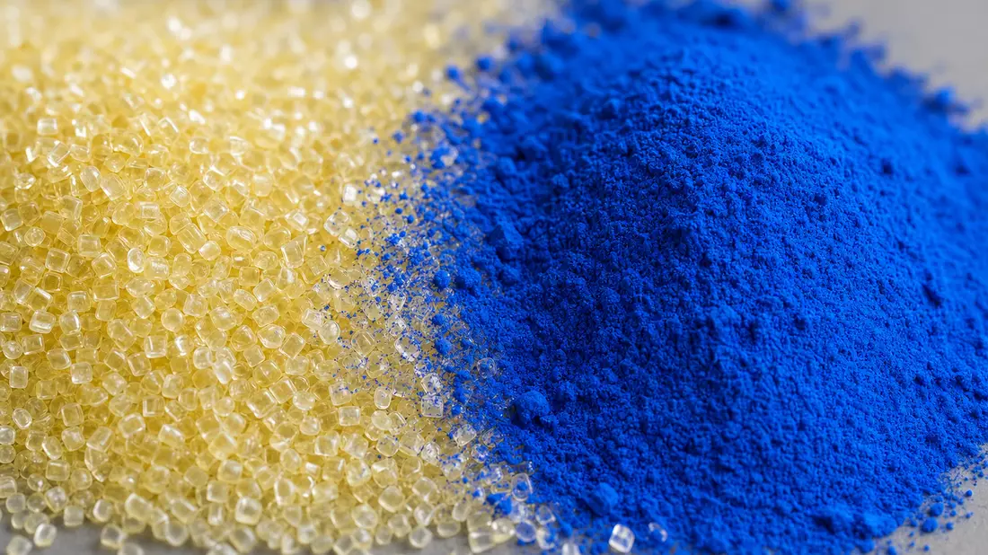 Makroaufnahme von transluzenten Polymer-Pellets neben vivid-blauem Pigmentpulver auf grauem Untergru