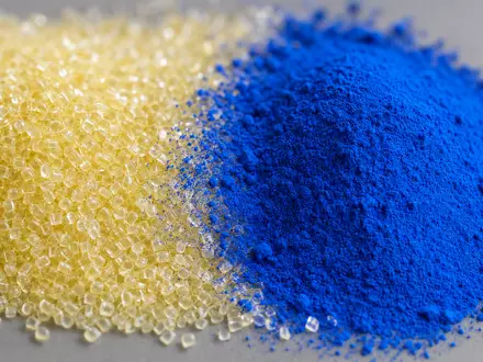 Extremes Makrofoto: Links ein Haufen transluzenter, blassgelblicher Polymer-Granulat-Pellets, rechts ein Häufchen leuchtendes Ultramarinblau-Pigmentpulver, beide durch einen feinen Übergangsstreifen getrennt. Auf neutralem Mittelgrau, aus leichter Vogelperspektive. Makroobjektiv, sehr geringe Schärfentiefe, sanfte Bokeh-Ränder. Weicher Softbox-Streiflicht von links oben, betont Textur und Granulat-Facetten - Foto: THN