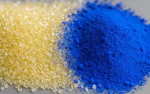Extremes Makrofoto: Links ein Haufen transluzenter, blassgelblicher Polymer-Granulat-Pellets, rechts ein Häufchen leuchtendes Ultramarinblau-Pigmentpulver, beide durch einen feinen Übergangsstreifen getrennt. Auf neutralem Mittelgrau, aus leichter Vogelperspektive. Makroobjektiv, sehr geringe Schärfentiefe, sanfte Bokeh-Ränder. Weicher Softbox-Streiflicht von links oben, betont Textur und Granulat-Facetten - Foto: THN