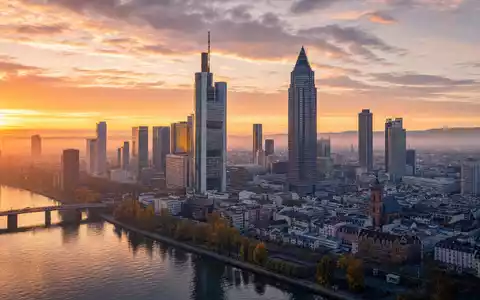 Fotorealistisches Luftbild des Frankfurter Bankenviertels bei Sonnenaufgang für Deutsche Bank AG (ISIN DE0005140008). Goldenes Morgenlicht trifft auf generische Hochhaustürme, der Main spiegelt die Skyline - Foto: THN