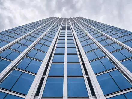 Architekturfoto eines generischen Glas-Stahl-Hochhauses von unten für Deutsche Bank AG (ISIN DE0005140008). Symmetrische Linien aus blauen Glasfassaden und weißen Stahlträgern konvergieren gen Himmel - Foto: THN