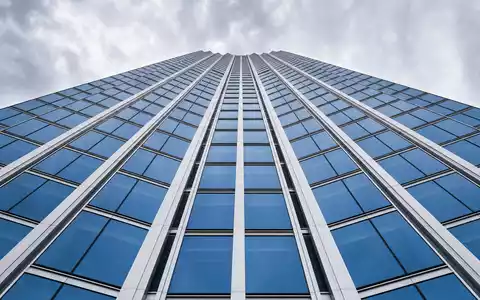 Architekturfoto eines generischen Glas-Stahl-Hochhauses von unten für Deutsche Bank AG (ISIN DE0005140008). Symmetrische Linien aus blauen Glasfassaden und weißen Stahlträgern konvergieren gen Himmel - Foto: THN