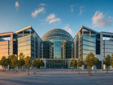 Architekturfoto im Stil der Deutsche Börse AG (ISIN DE0005810055): Modernes europäisches Finanzdistrikt-Gebäude mit markanter Glaskuppel, symmetrischer Fassade aus Glas und Stahl und beleuchtetem Platz bei goldenem Abendlicht - Foto: THN
