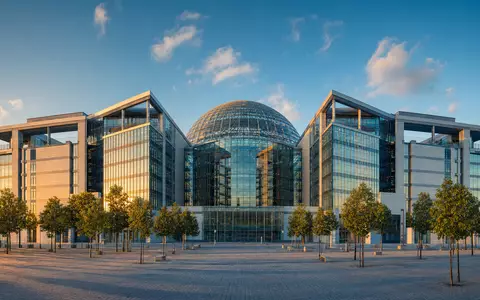 Architekturfoto im Stil der Deutsche Börse AG (ISIN DE0005810055): Modernes europäisches Finanzdistrikt-Gebäude mit markanter Glaskuppel, symmetrischer Fassade aus Glas und Stahl und beleuchtetem Platz bei goldenem Abendlicht - Foto: THN