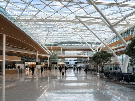Architekturaufnahme für Deutsche Lufthansa AG (ISIN DE0008232125): Moderne Flughafen-Terminalhalle mit großem Glasdach aus Stahlfachwerkstruktur, helles Tageslicht fällt von oben ein, silhouettierte Passagiere auf dem Concourse - Foto: THN