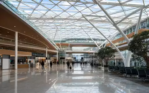 Architekturaufnahme für Deutsche Lufthansa AG (ISIN DE0008232125): Moderne Flughafen-Terminalhalle mit großem Glasdach aus Stahlfachwerkstruktur, helles Tageslicht fällt von oben ein, silhouettierte Passagiere auf dem Concourse - Foto: THN