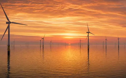 Fotorealistisches Panorama eines Offshore-Windparks in der Nordsee bei goldenem Sonnenuntergang. Zahlreiche weiße Windturbinen stehen im ruhigen Wasser, der orangefarbene Himmel spiegelt sich auf der glatten Meeresoberfläche. Dieses Bild steht exemplarisch für die Offshore-Windkraft-Aktivitäten von E.ON SE (ISIN DE000ENAG999) im Bereich erneuerbare Energien - Foto: THN