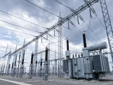 Architektonische Aufnahme eines modernen Umspannwerks mit imposantem Stahlgittergerüst, Porzellan- und Polymerisolatoren sowie massiven Transformatoren. Die Perspektive von unten betont die industrielle Präzision der Hochspannungsinfrastruktur vor bewölktem Himmel. Dieses Bild repräsentiert die Kernkompetenz von E.ON SE (ISIN DE000ENAG999) im Bereich Stromnetze - Foto: THN