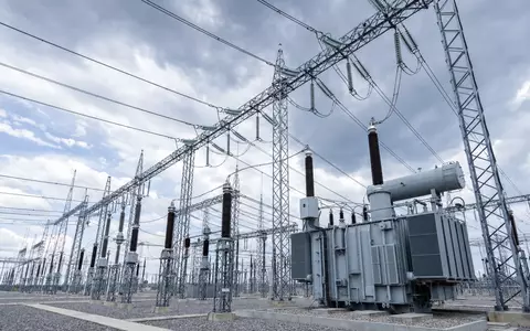 Architektonische Aufnahme eines modernen Umspannwerks mit imposantem Stahlgittergerüst, Porzellan- und Polymerisolatoren sowie massiven Transformatoren. Die Perspektive von unten betont die industrielle Präzision der Hochspannungsinfrastruktur vor bewölktem Himmel. Dieses Bild repräsentiert die Kernkompetenz von E.ON SE (ISIN DE000ENAG999) im Bereich Stromnetze - Foto: THN
