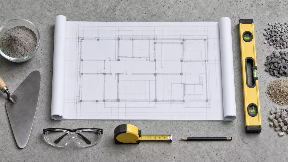 Flatlay: Bauplan, Zementprobe, Maurerkelle, Wasserwaage und Schutzbrille auf Betonuntergrund