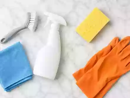 Aufgeräumte Draufsicht-Produktfotografie von generischen Reinigungsmitteln auf weißer Marmoroberfläche: Sprühflasche, Scheuerbürste, blaues Mikrofasertuch, gelber Schwamm und orangefarbene Gummihandschuhe – repräsentiert das Reinigungs- und Haushaltsproduktportfolio der Henkel AG & Co. KGaA (ISIN DE0006048432) - Foto: THN