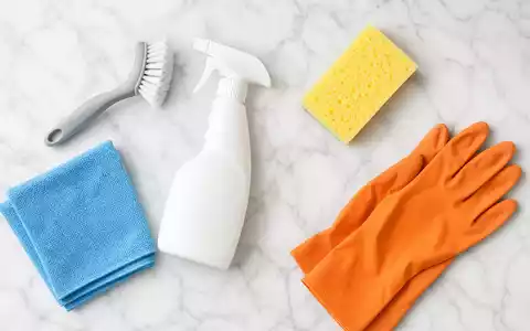 Aufgeräumte Draufsicht-Produktfotografie von generischen Reinigungsmitteln auf weißer Marmoroberfläche: Sprühflasche, Scheuerbürste, blaues Mikrofasertuch, gelber Schwamm und orangefarbene Gummihandschuhe – repräsentiert das Reinigungs- und Haushaltsproduktportfolio der Henkel AG & Co. KGaA (ISIN DE0006048432) - Foto: THN