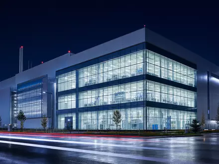 Architektonisches Nachtfoto eines modernen Halbleiterwerks mit leuchtenden Reinraumfenstern – vergleichbar mit den Produktionsstandorten von Infineon Technologies AG (ISIN DE0006231004) in der Region München und weltweit - Foto: THN