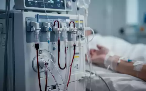 Nahaufnahme einer Dialysemaschine mit Schläuchen und einem leicht unscharfen Patientenarm im Hintergrund, der eine Nadel zeigt. - Foto: über boerse-global.de