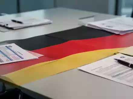 Nahaufnahme einer stilisierten deutschen Flagge auf einem Konferenztisch mit unscharfen Dokumenten und Stift, symbolisiert Verhandlungen. - Foto: über boerse-global.de