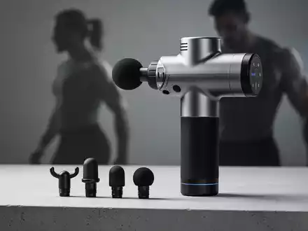 Eine moderne Perkussionsmassagepistole mit verschiedenen Aufsätzen auf einer strukturierten Oberfläche, im Hintergrund eine Person. - Foto: über boerse-global.de