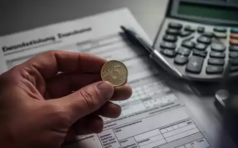 Eine Hand hält eine kleine Münze vor einem unscharfen Hintergrund mit Finanzdokumenten und einem Taschenrechner. - Foto: über boerse-global.de