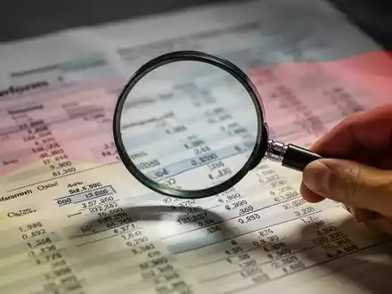 Eine Hand hält eine Lupe über ein komplexes Finanzdokument mit verschwommenem Text und Zahlen, das Steuerausfälle symbolisiert. - Foto: über boerse-global.de
