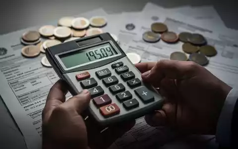 Nahaufnahme einer Hand, die einen Taschenrechner mit Zahlen hält, mit unscharfen Euro-Münzen und Dokumenten im Hintergrund. - Foto: über boerse-global.de