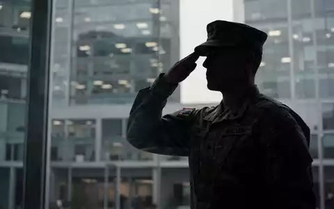 Ein Soldat in Uniform salutiert vor einem unscharfen Hintergrund eines modernen Bürogebäudes, der die Verbindung von Militär und Zivilberuf darstellt. - Foto: über boerse-global.de