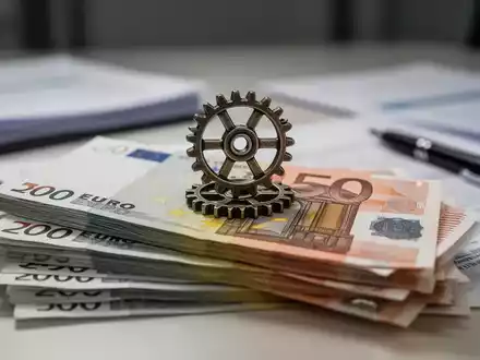 Ein Stapel Euro-Banknoten mit einem kleinen Zahnrad darauf, im Hintergrund verschwommene Büroumgebung. Symbolisiert kleine Reformen. - Foto: über boerse-global.de