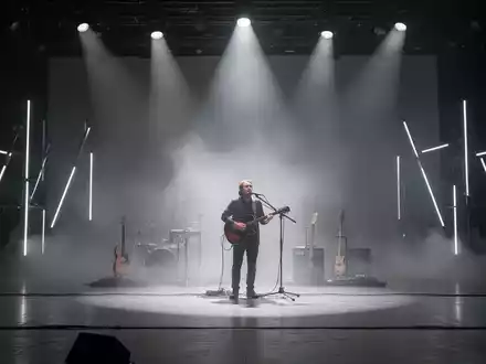 Ein einzelner Musiker auf einer spärlich beleuchteten Bühne, umgeben von Nebel, symbolisiert Minimalismus in der Musik. - Foto: über boerse-global.de