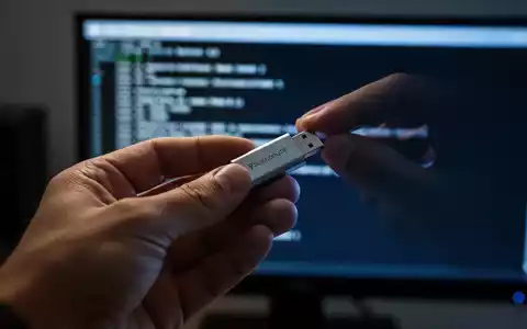 Eine Hand hält einen USB-Stick, der im Licht eines Computerbildschirms mit technischen Informationen leuchtet. - Foto: über boerse-global.de
