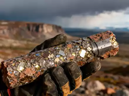 Nahaufnahme einer Bohrkernprobe mit metallischen Sprenkeln, gehalten von einer behandschuhten Hand vor einer rauen Landschaft. - Foto: über boerse-global.de