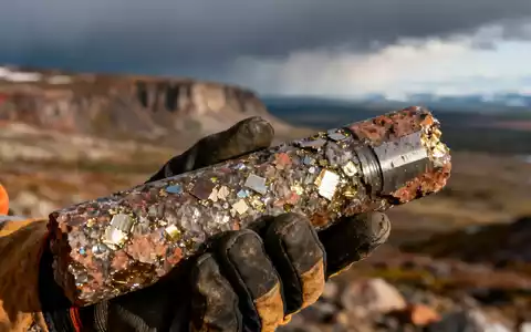 Nahaufnahme einer Bohrkernprobe mit metallischen Sprenkeln, gehalten von einer behandschuhten Hand vor einer rauen Landschaft. - Foto: über boerse-global.de